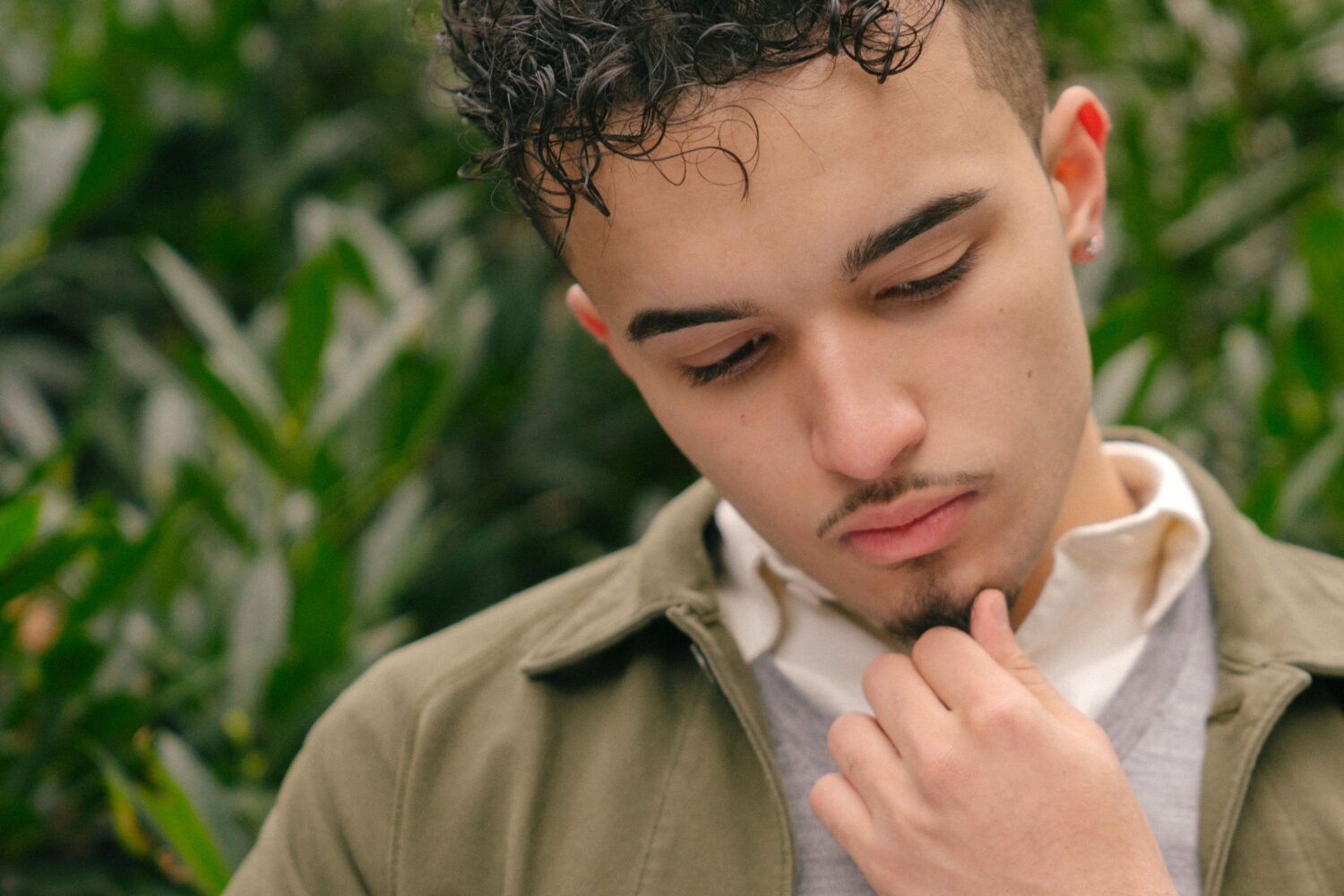 Young person in an olive jacket looking down thoughtfully, hand near chin, against leafy greenery.