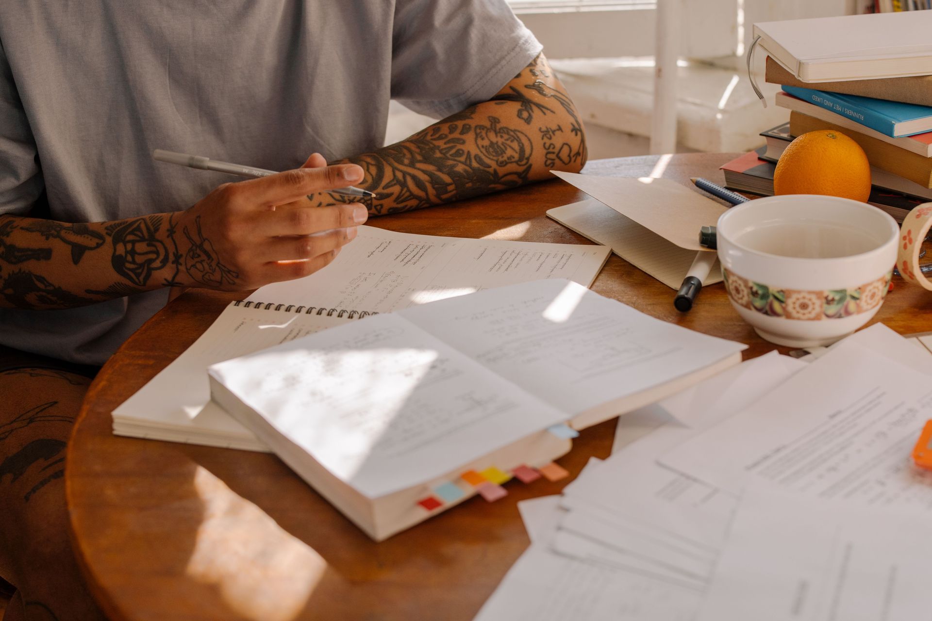 Person writing at a sunlit table with papers, notebooks, a mug, and an orange nearby.