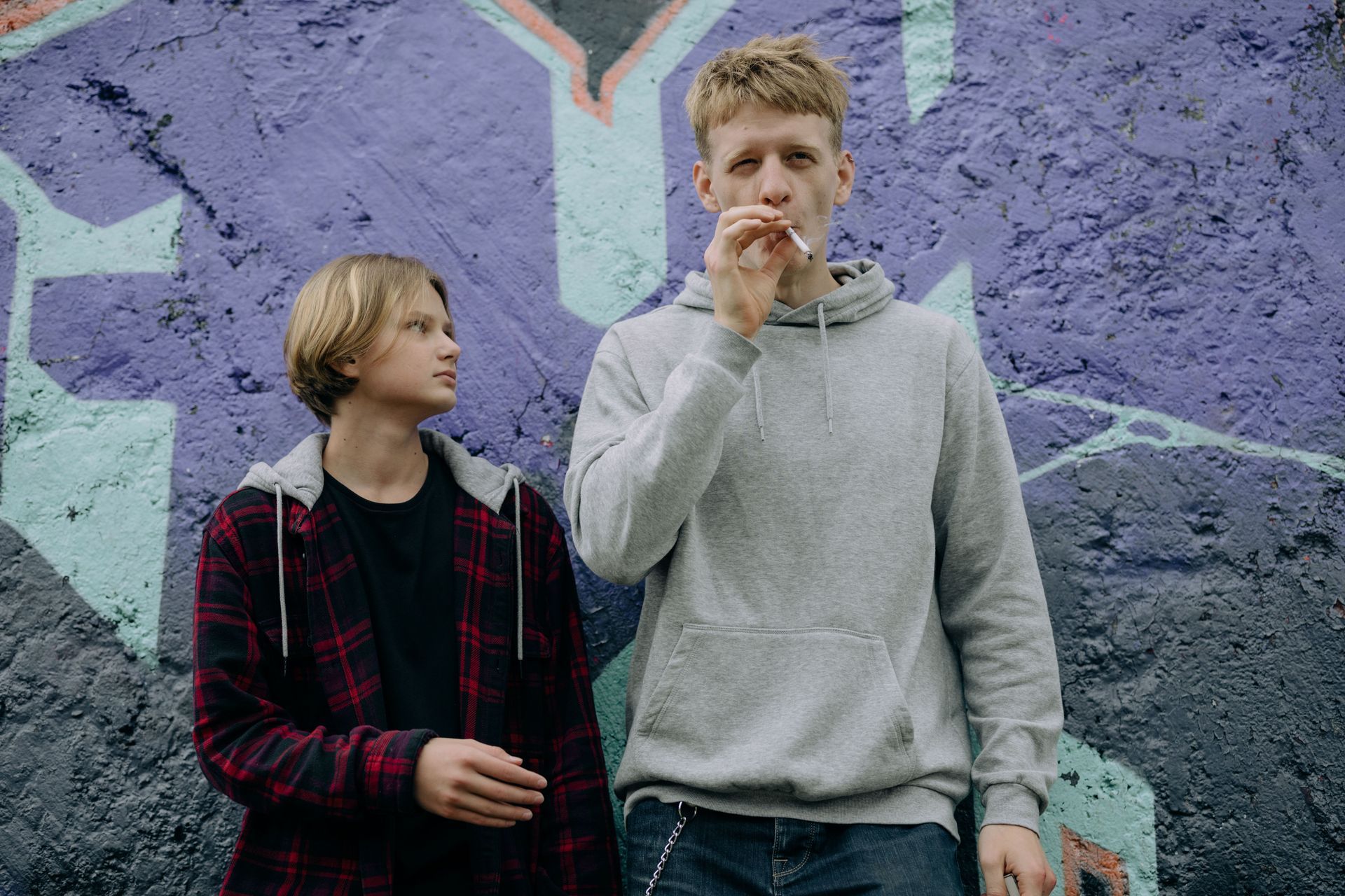 Two people standing by a purple graffiti wall, one eating a snack and the other looking at them.