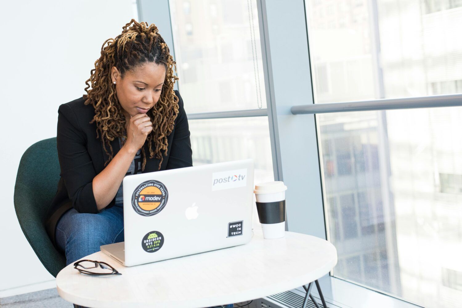 Person working on a laptop at a white table in a bright office lounge, with a coffee cup nearby