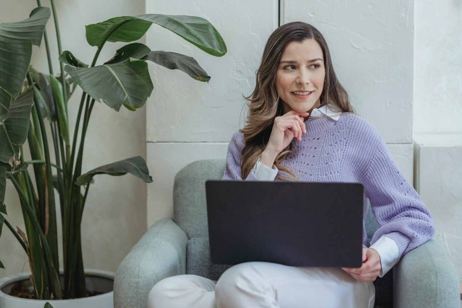 Woman sitting on a gray couch with a laptop, smiling in a bright room with large green plants