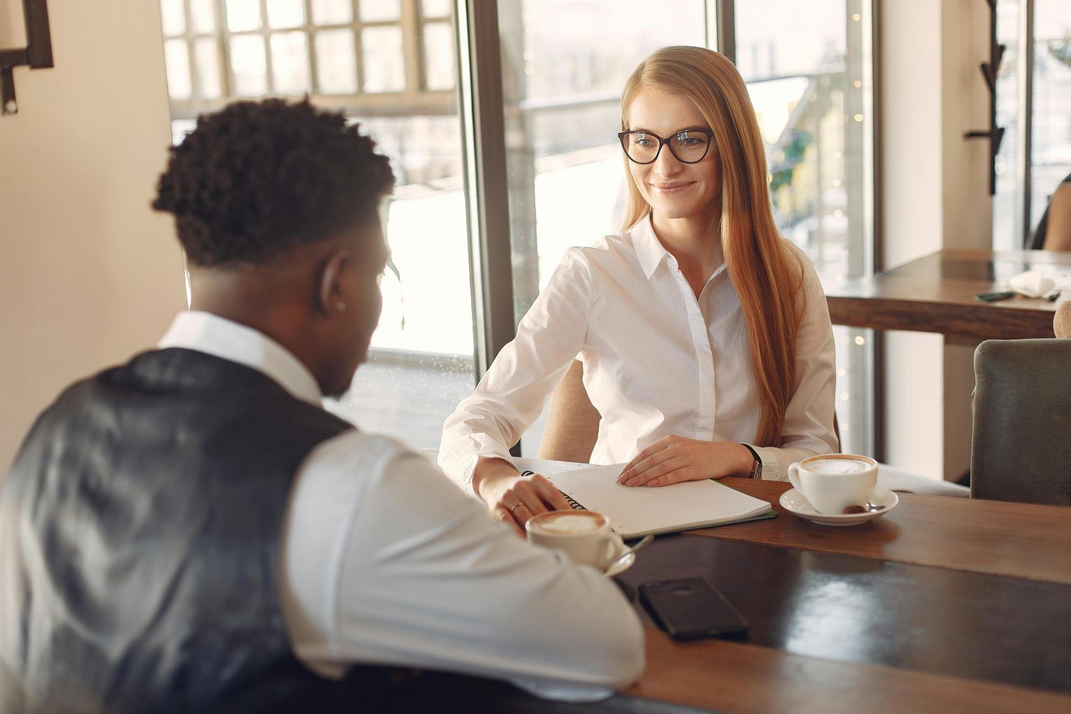 Two people in a sunlit office discuss documents across a table with coffee cups.