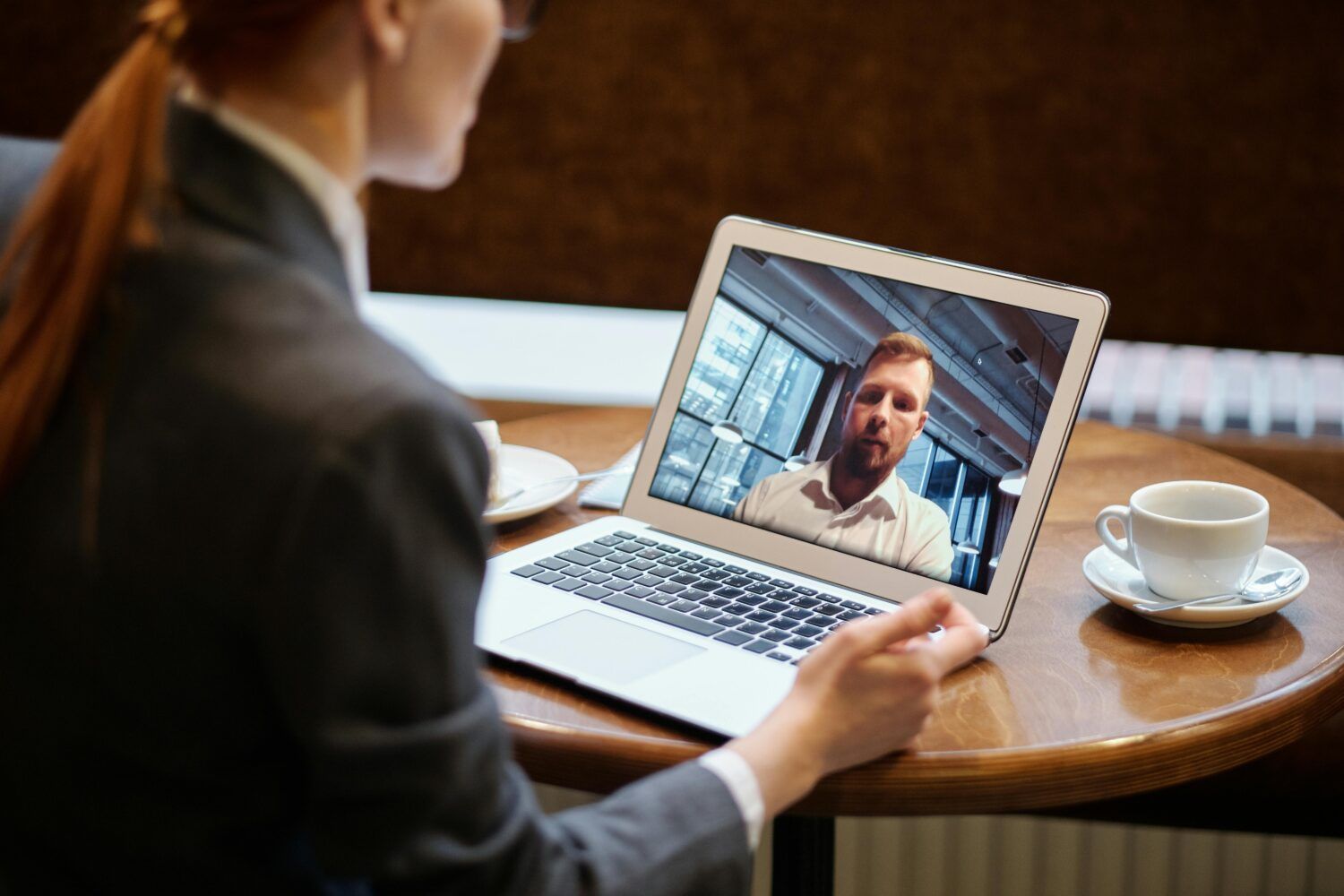 Woman in a blazer video chatting on a laptop at a café table with a cup of coffee.