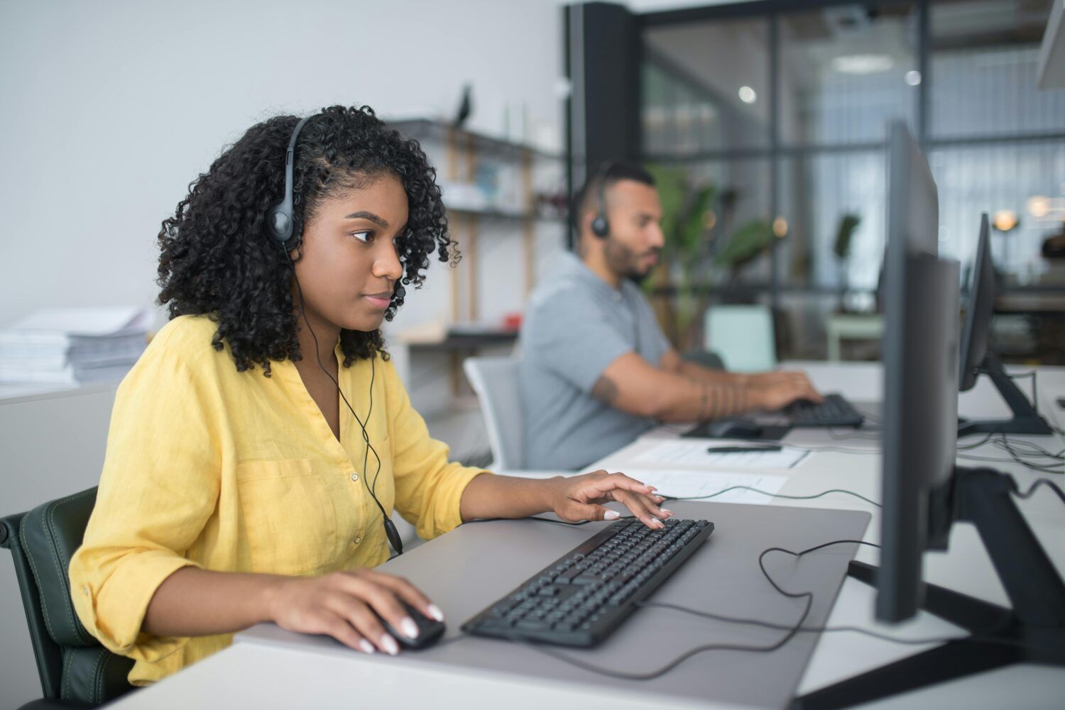 Person typing at a desktop computer in an office workspace with other employees in the background