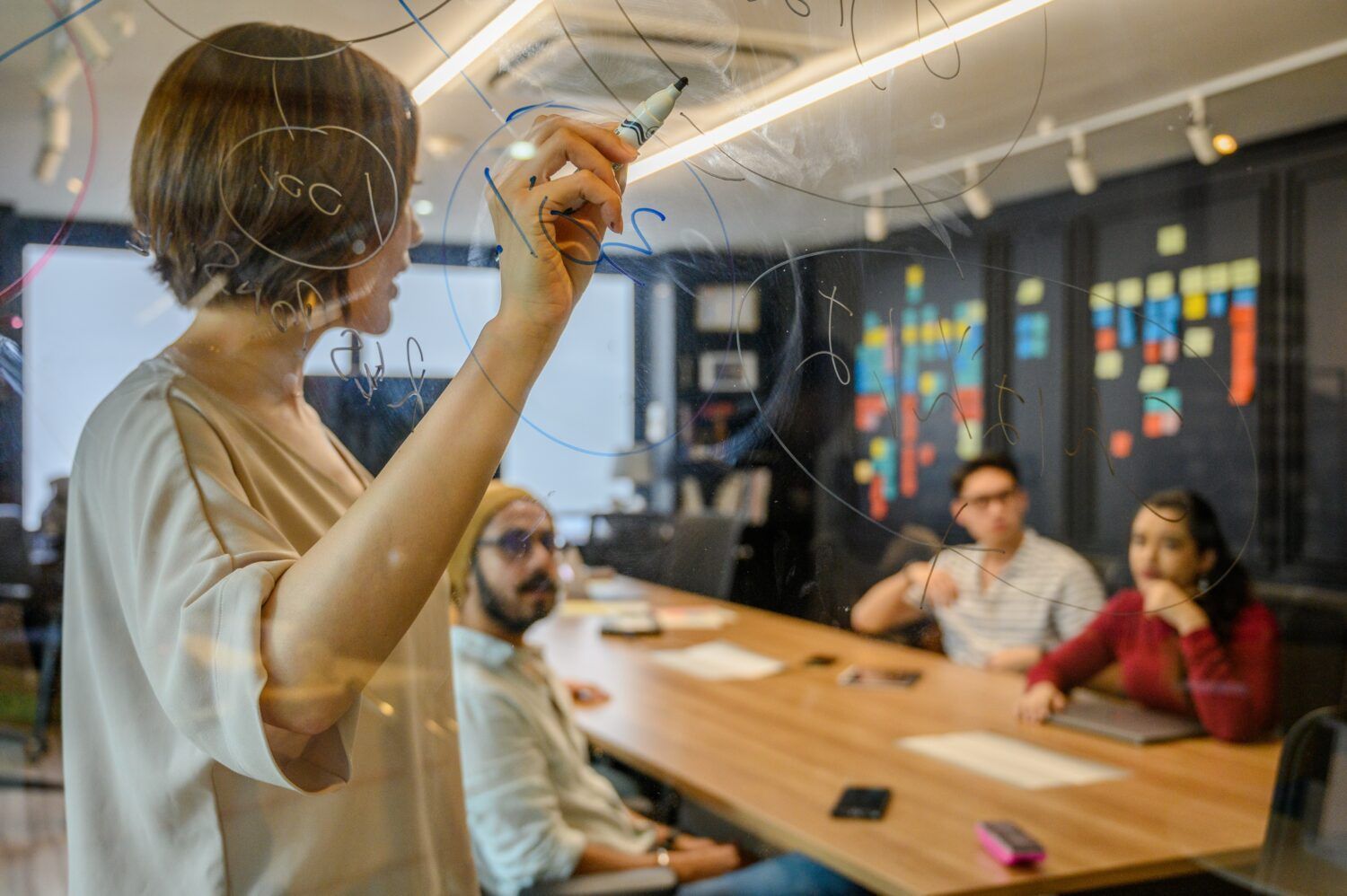 Person writing on a glass wall in a meeting room while others sit at a conference table
