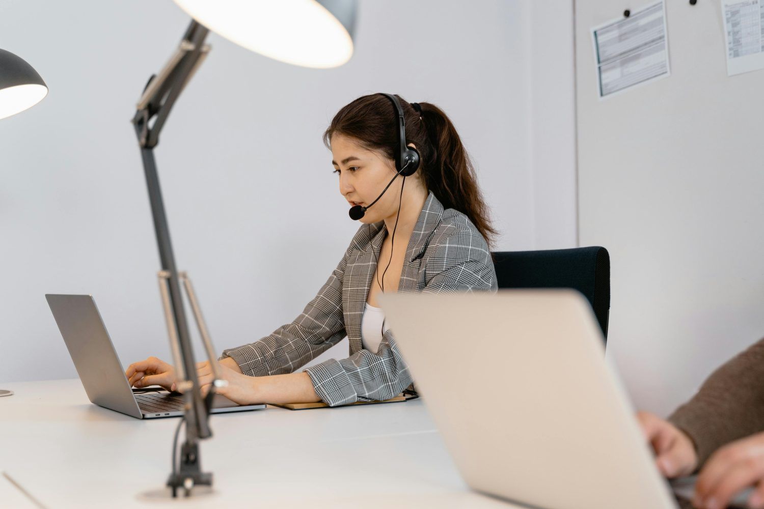 Woman wearing headset working on a laptop at a white office desk under a lamp