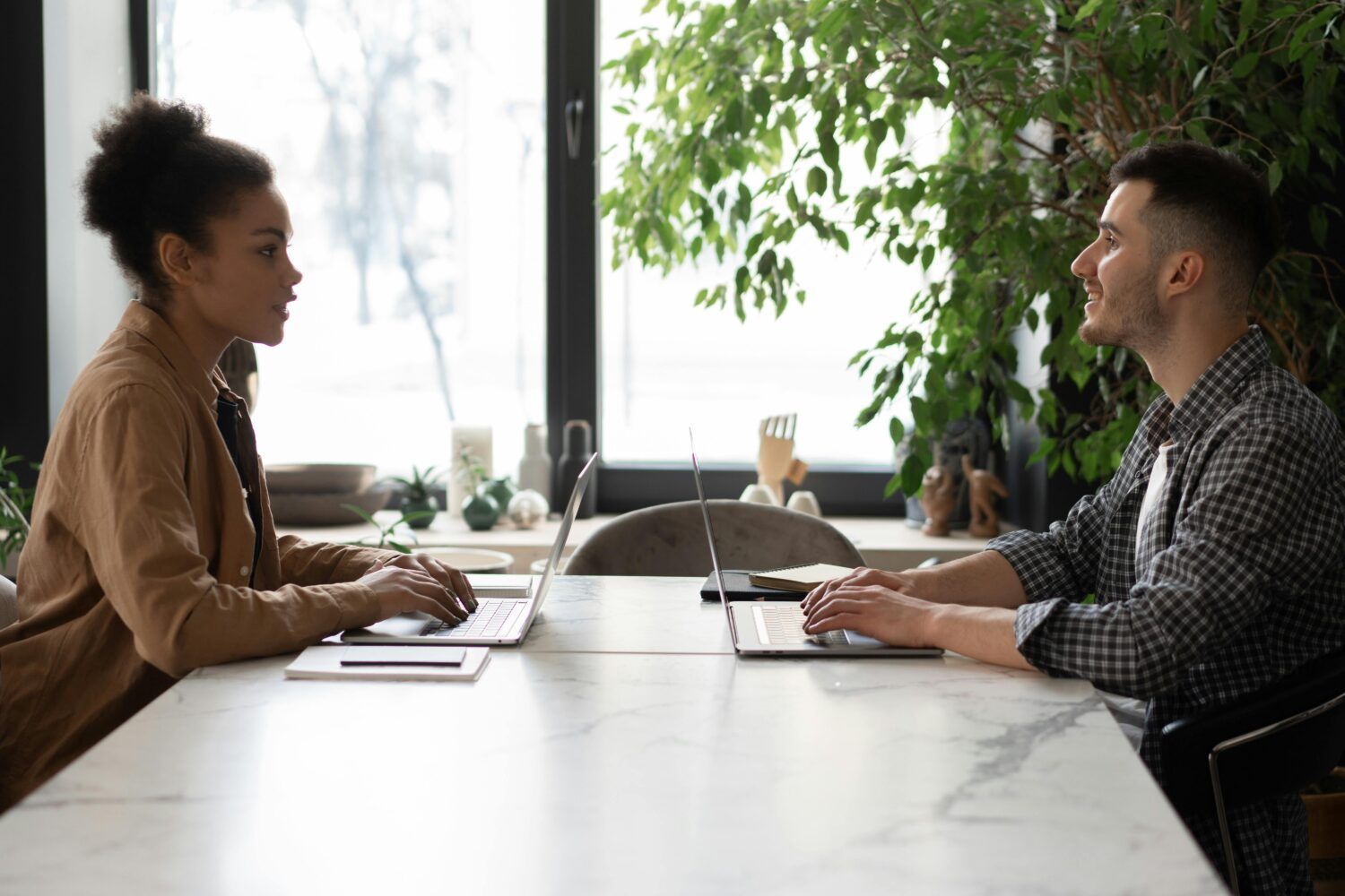 Two people sitting across a table in a bright office, working on laptops during a meeting.