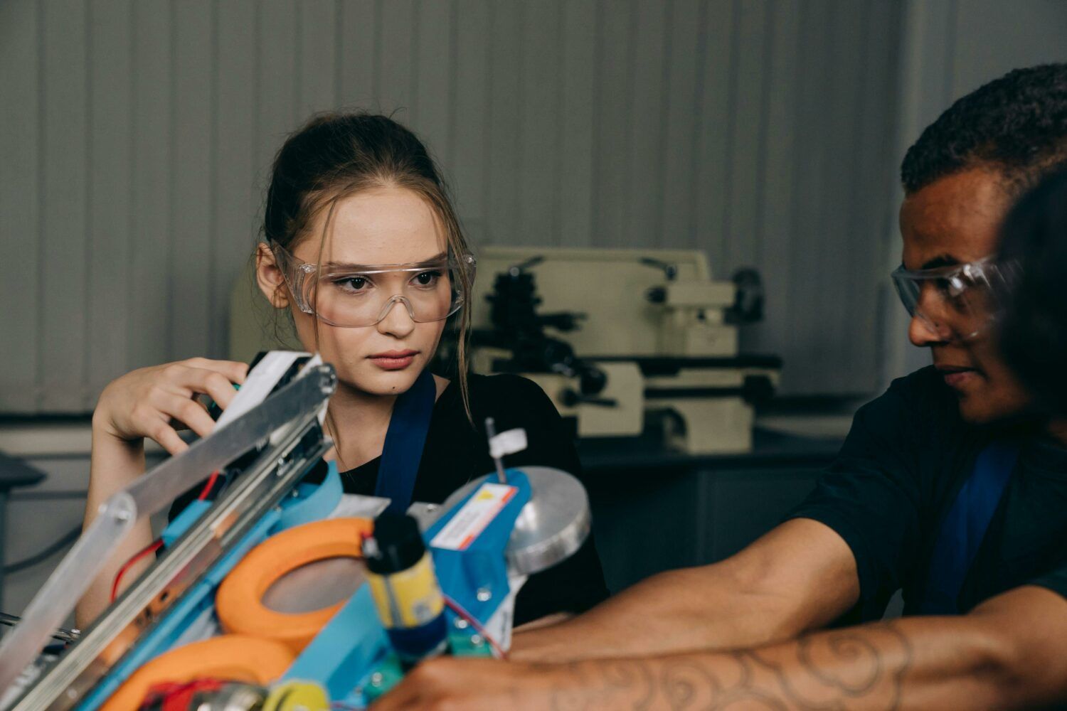 Two people wearing safety glasses work with tools at a workbench in a workshop.