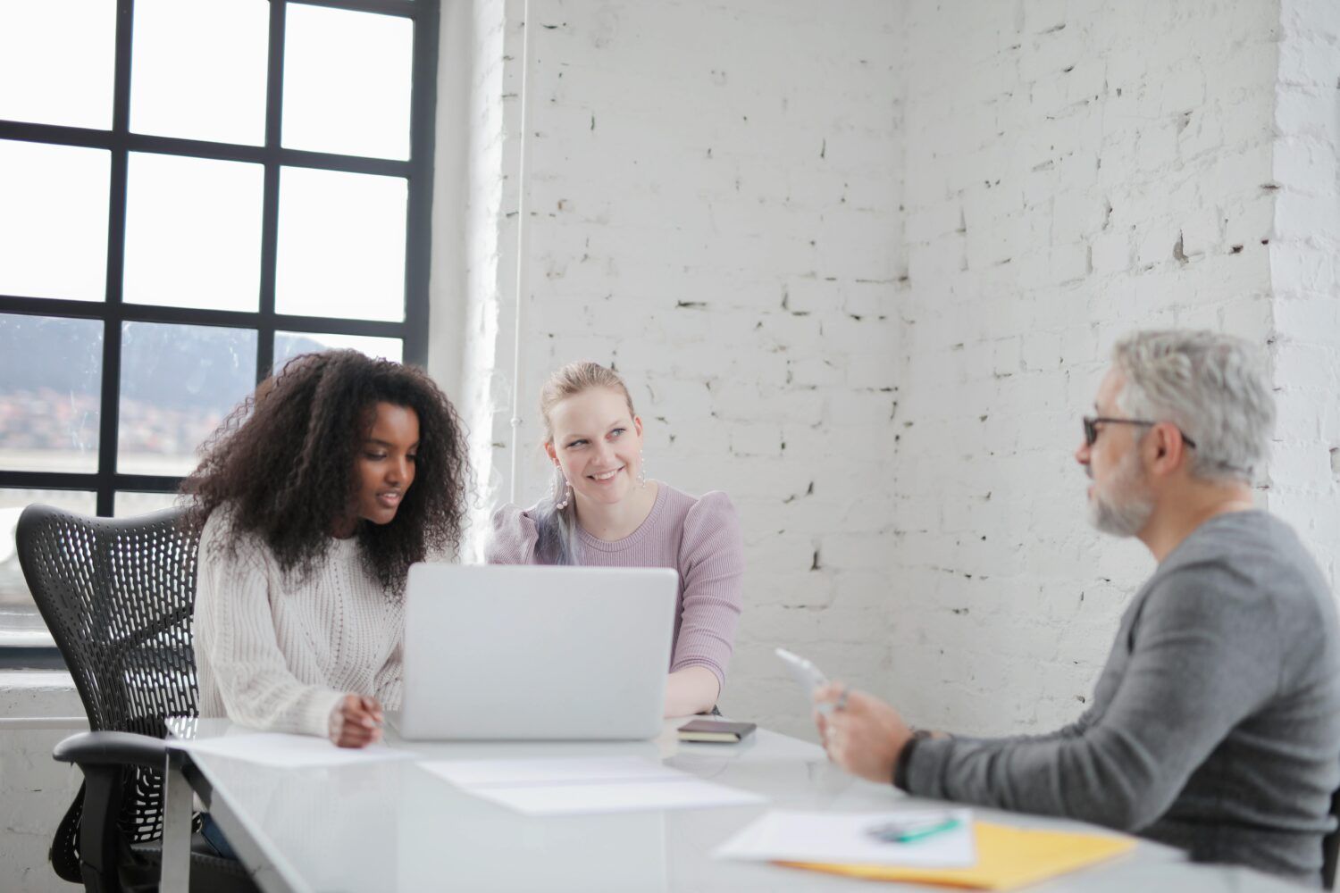Three people in a bright office meeting around a laptop at a white table.