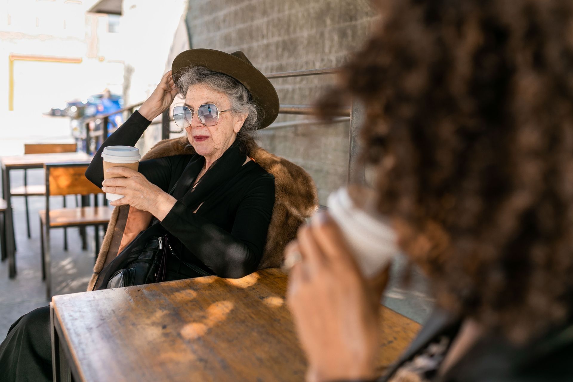 Woman in a hat and sunglasses holds a drink at a café table, seen past another blurred diner.