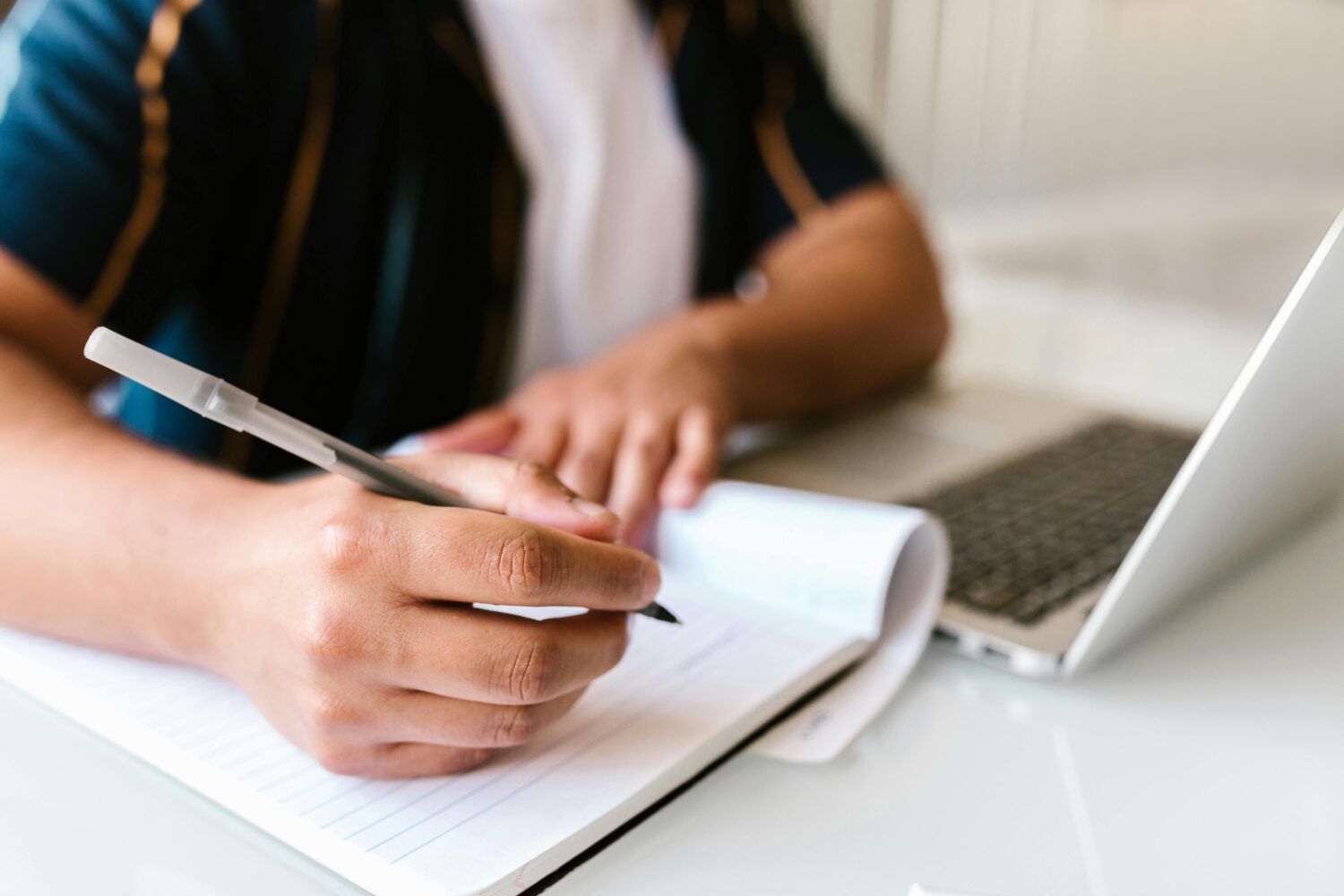 Hand writing notes in a notebook beside a silver laptop on a desk