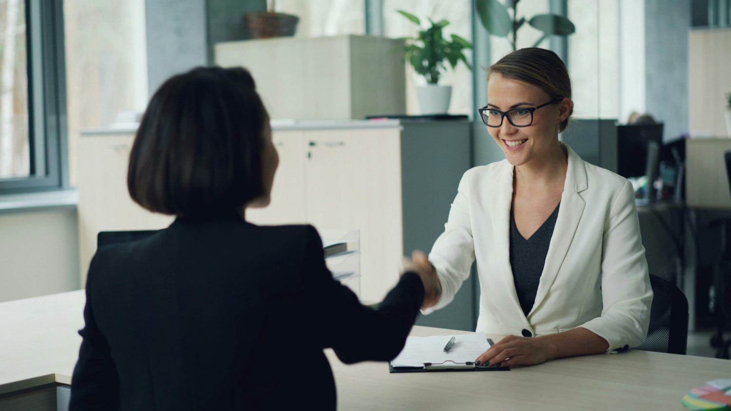 Two people shaking hands across a desk in a bright office meeting room.