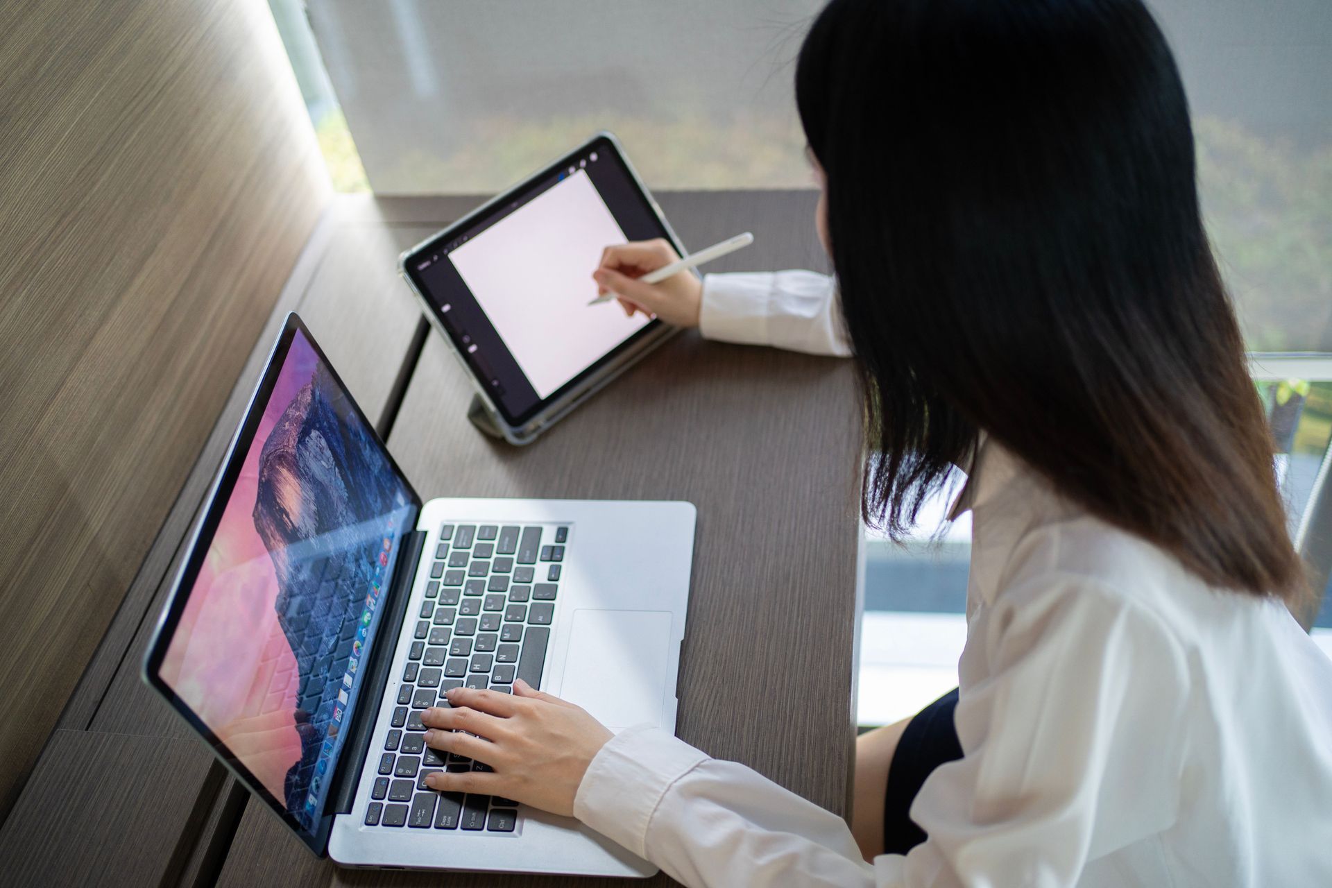 Person working on a laptop while using a tablet on a desk in a narrow workspace