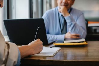 Two people in a meeting, one taking notes beside a laptop and notebook on a wooden table.