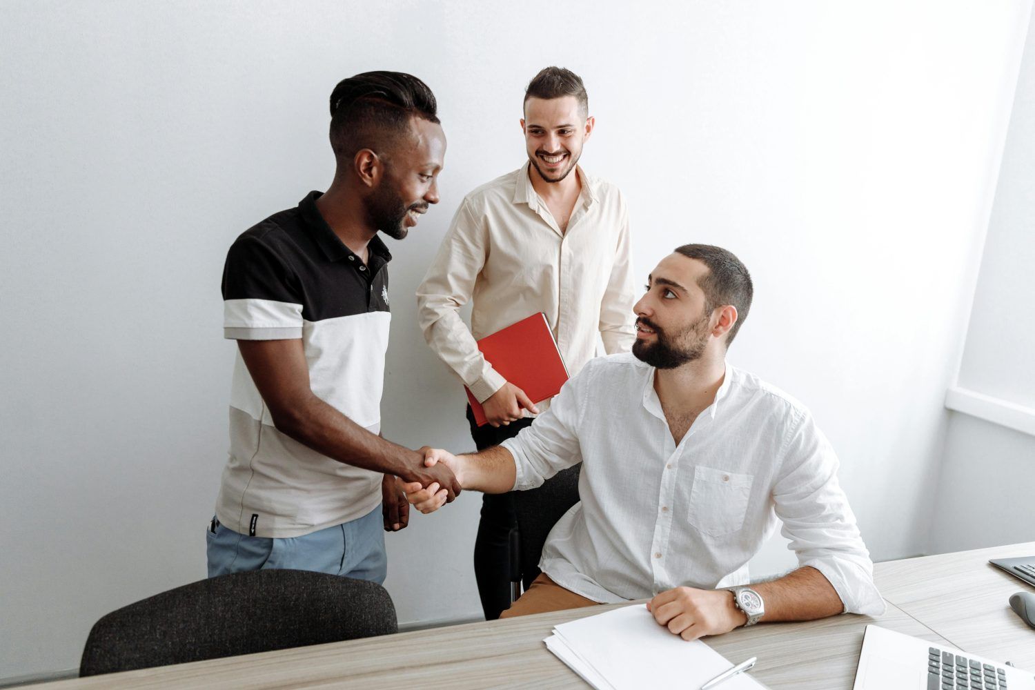 Two colleagues shake hands while a third stands behind holding a red folder in a bright office.