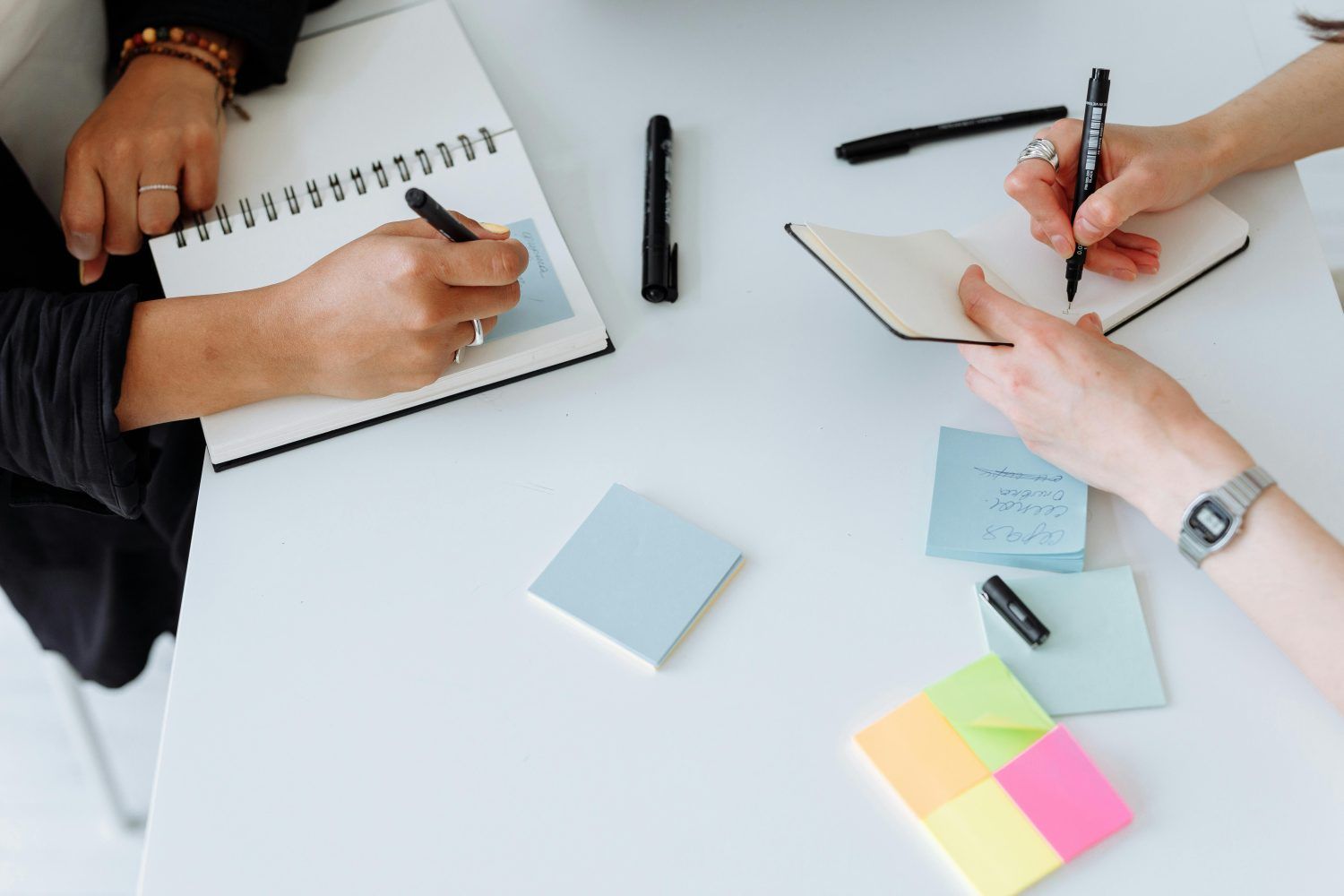 Two people writing in notebooks at a white table with sticky notes and pens.