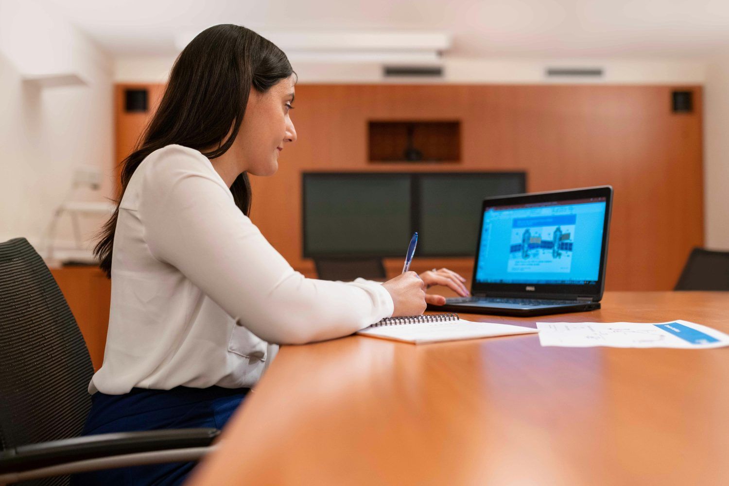 Woman writing at a conference table beside an open laptop and papers in a meeting room