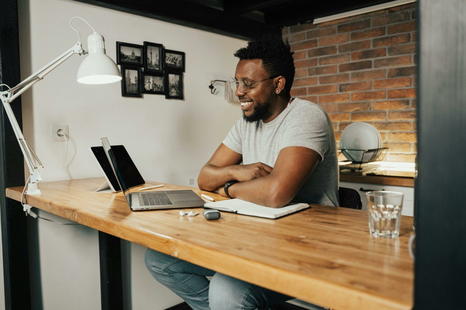 Man smiling at a desk with a laptop and notebook in a brick-walled home office