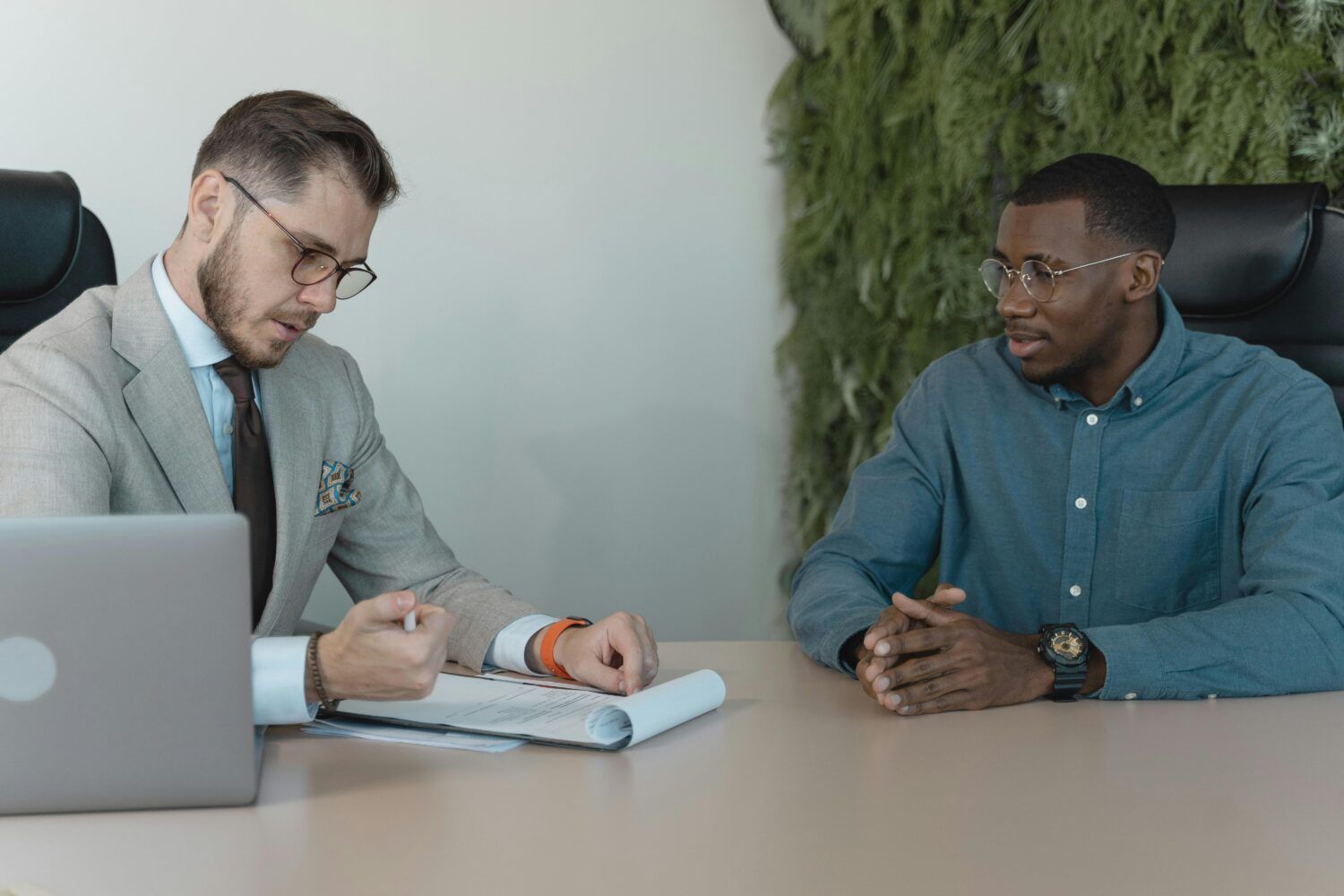 Two people in a meeting at a desk, discussing notes beside a laptop.