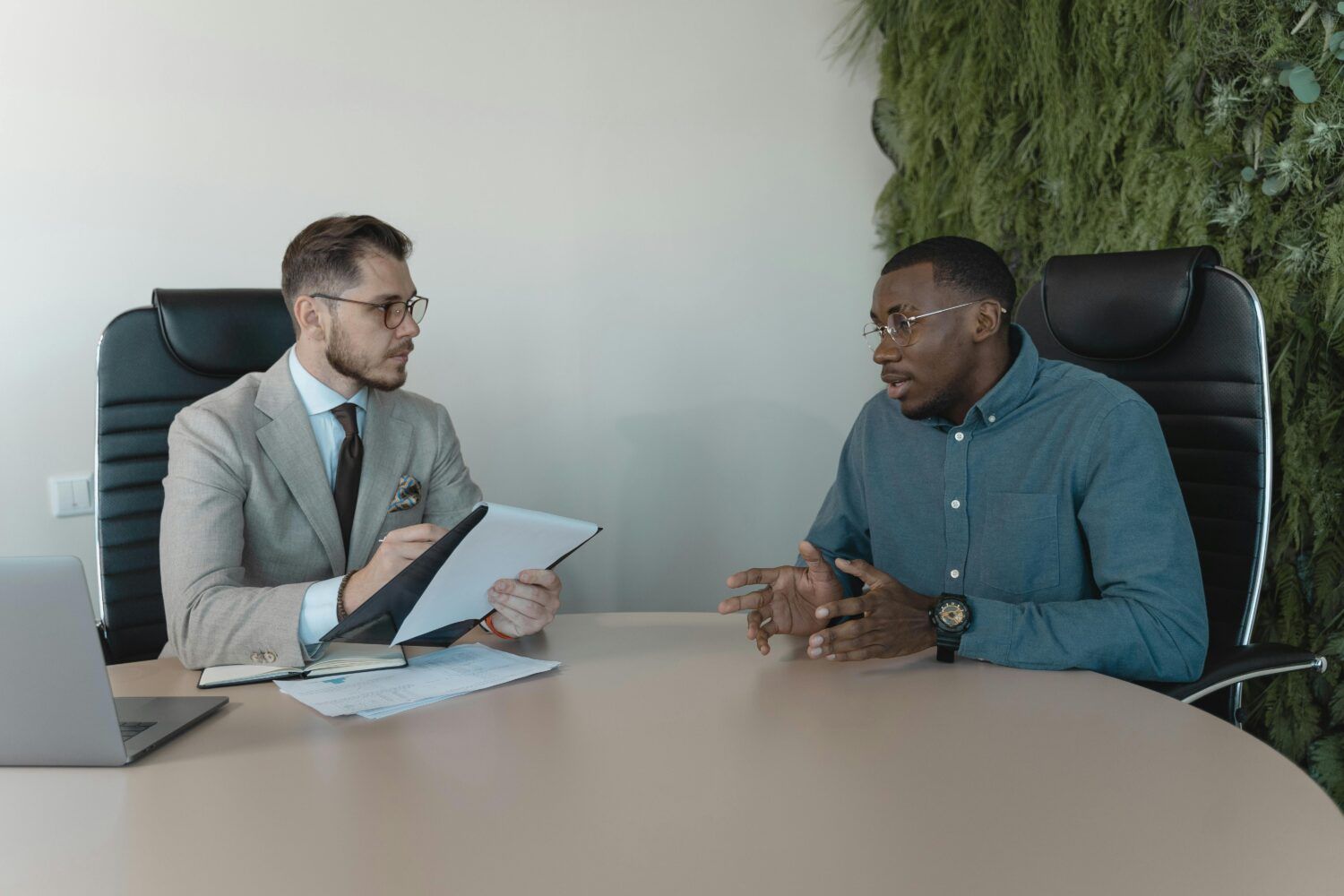 Two people in an office meeting, one holding papers while talking across a conference table.