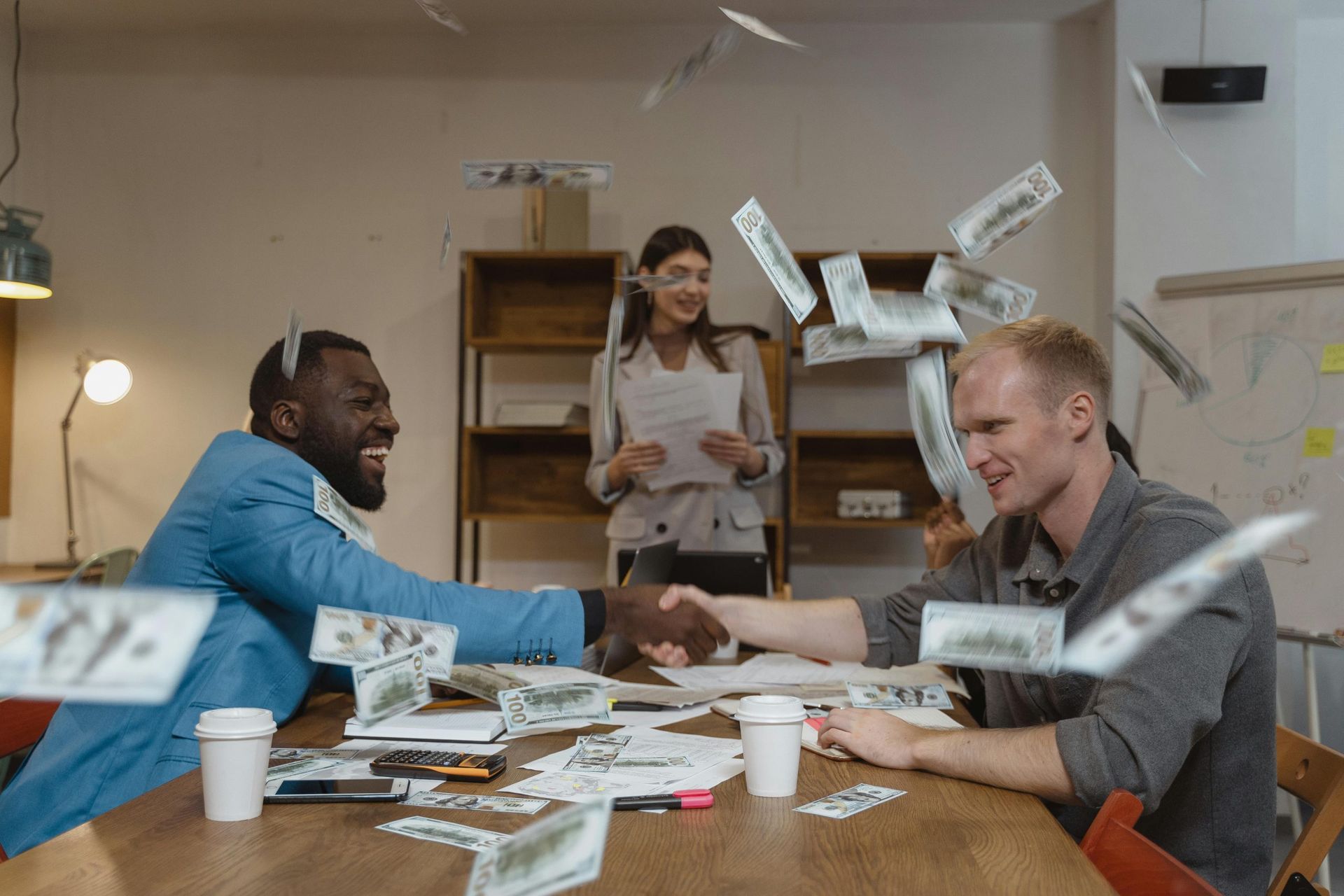 Two people at a table smiling as cash flies around them during a lively office celebration