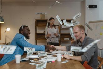 Two people at a table smiling as cash flies around them during a lively office celebration
