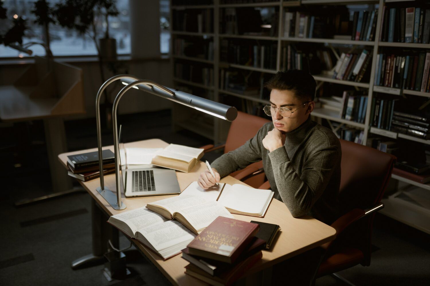 Person studying at a desk with books and laptop in a dim library, under a lamp