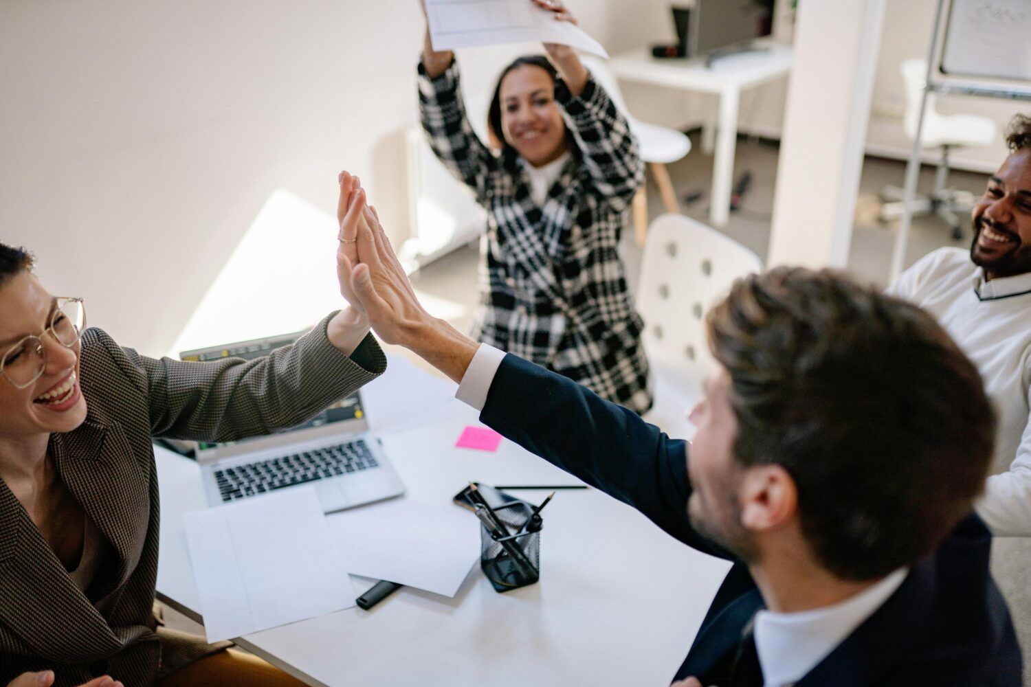 Coworkers in a meeting high-five over a table with papers and a laptop