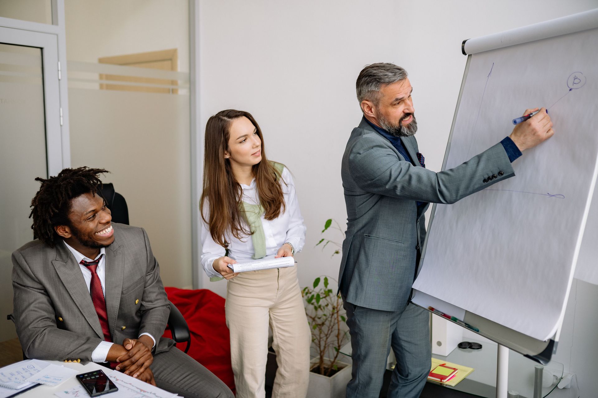 Three coworkers in a meeting room, one writing on a flip chart while two listen nearby.