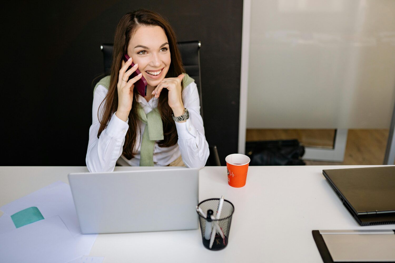 Woman on the phone at a desk with a laptop, smiling in a modern office