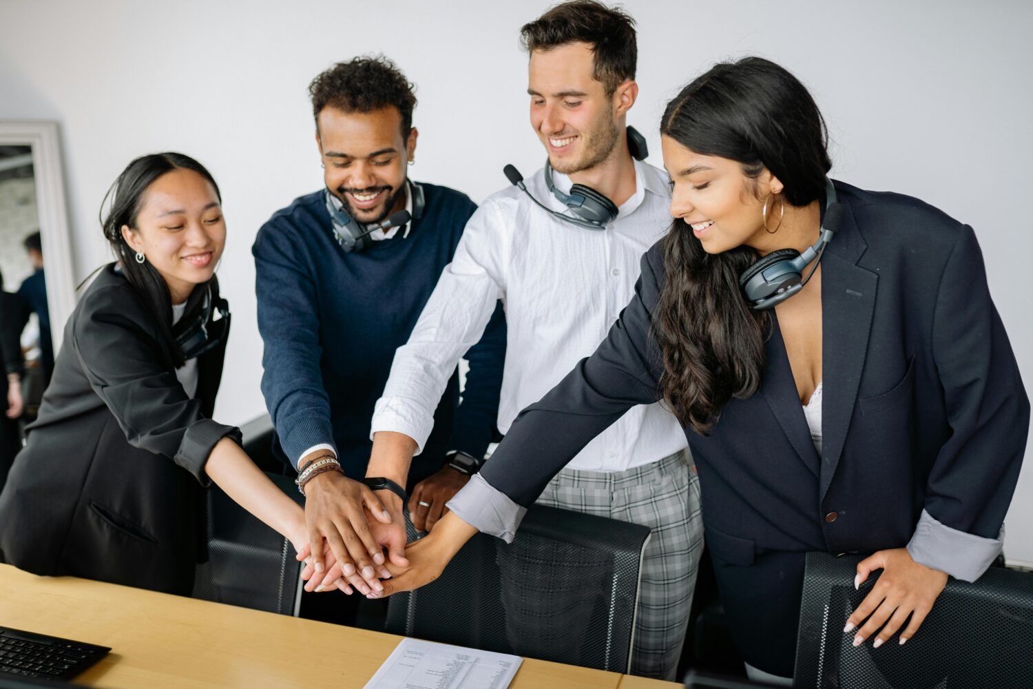 Four coworkers stack hands together in an office, smiling in a team huddle.