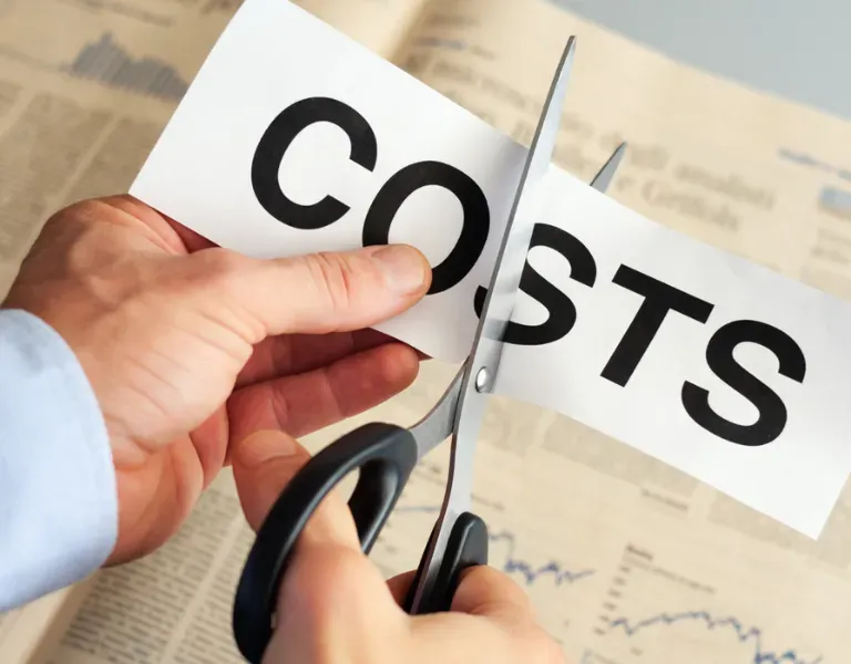 Hands cutting a paper labeled “COSTS” with scissors over a financial chart background