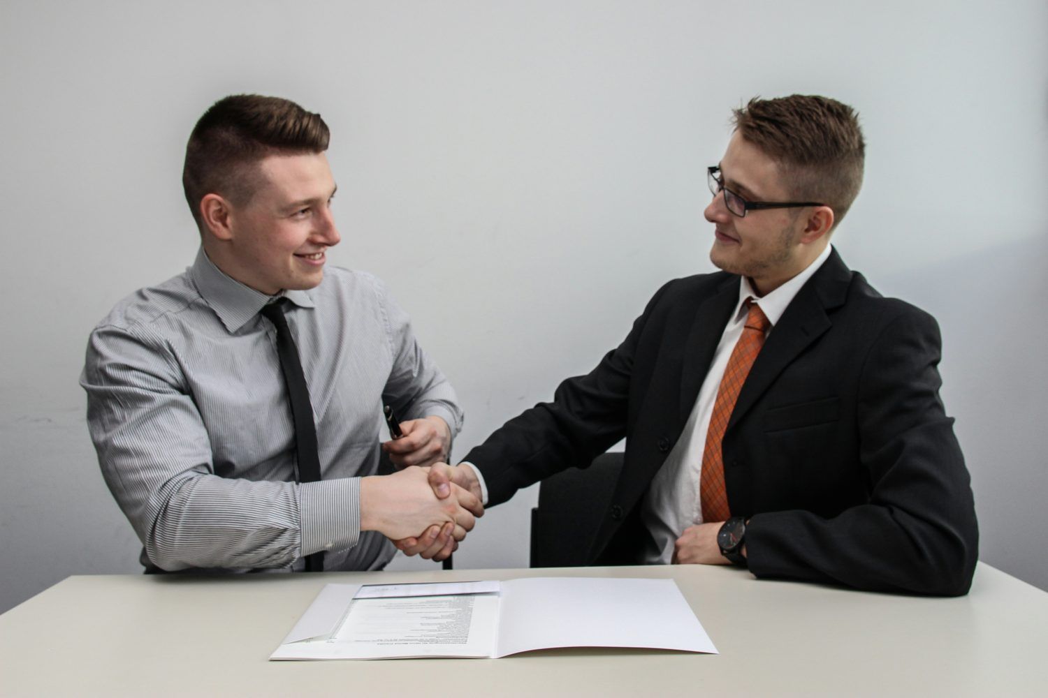 Two men in business attire shaking hands across a table with papers on it