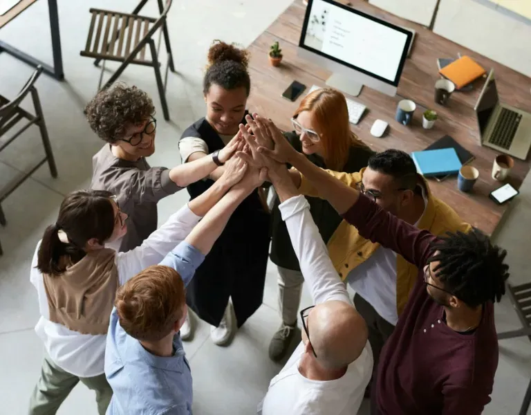 Coworkers in a meeting huddle, stacking hands in a team huddle around a desk with a computer monitor.