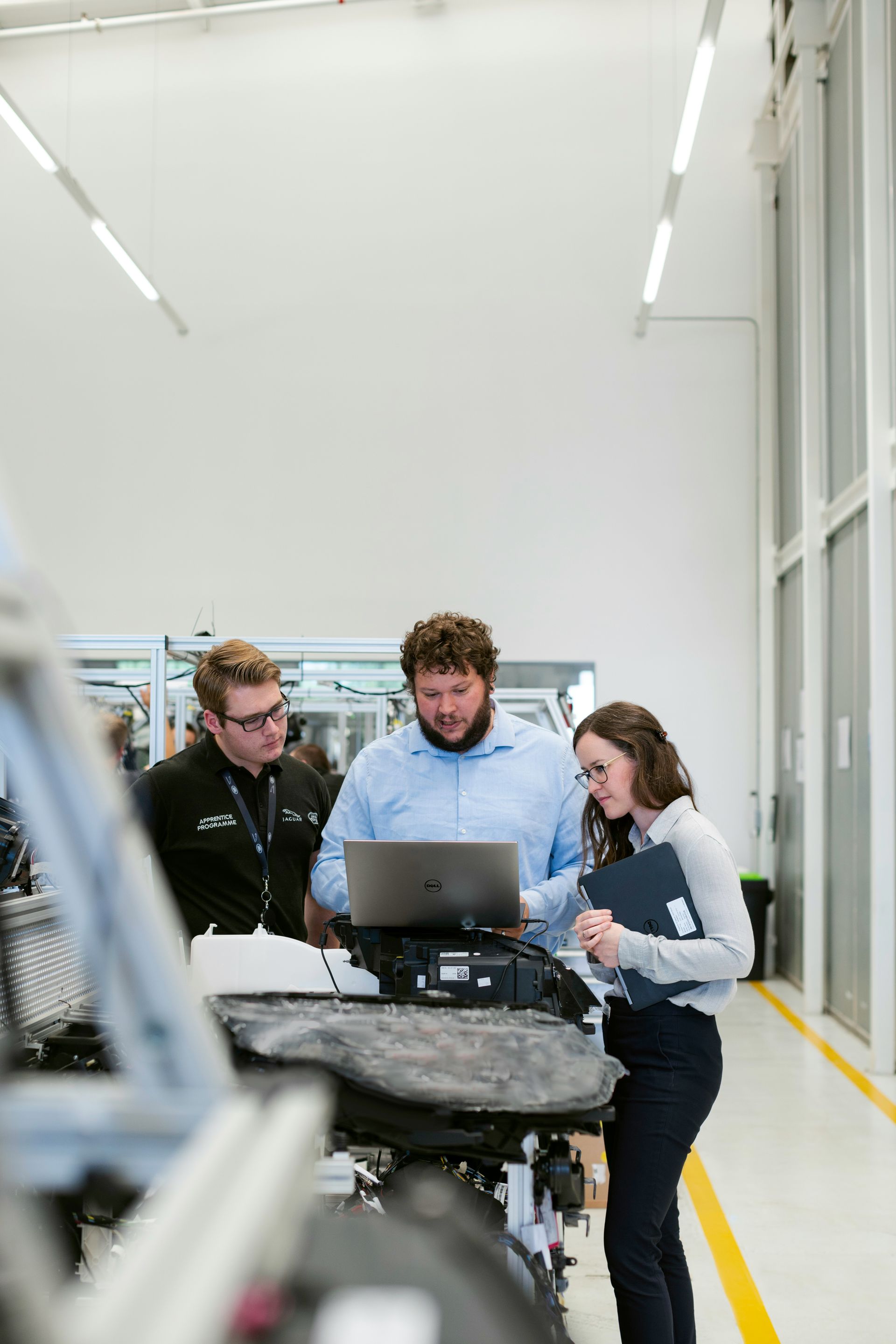 Three people gather around a laptop in a bright industrial workspace, examining machinery.