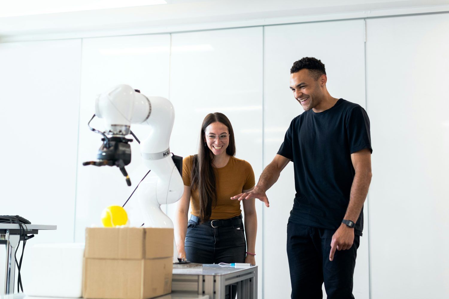 Two people in a bright lab stand beside a white robot arm placing a yellow object into a cardboard box.