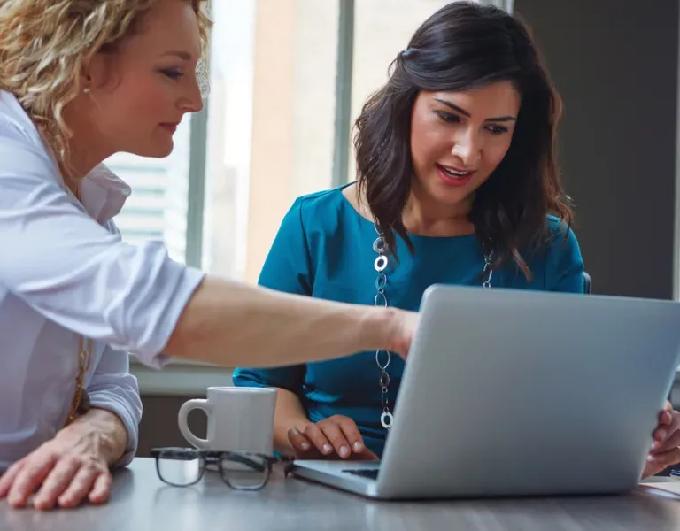 Two coworkers review a laptop at a table, with one pointing at the screen and a coffee mug nearby.