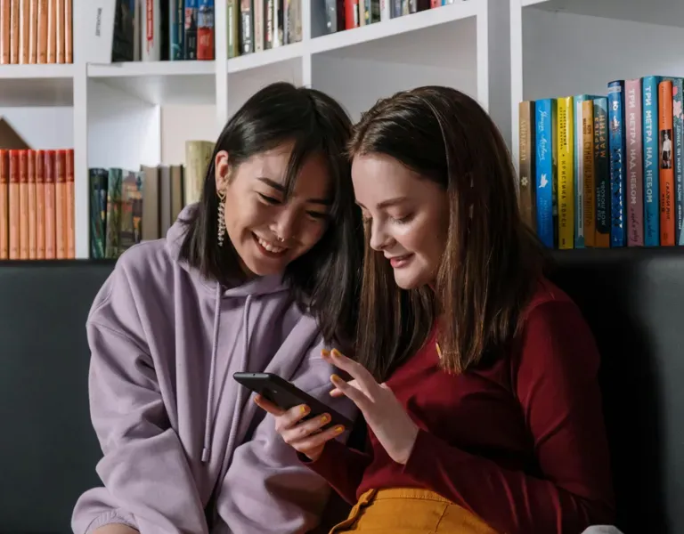Two people smiling on a couch, looking at a phone in a cozy room with bookshelves.