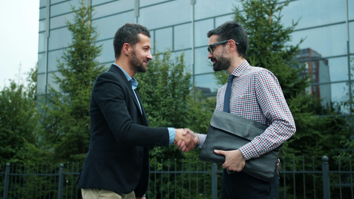 Two men shake hands outside an office building, one holding a folder