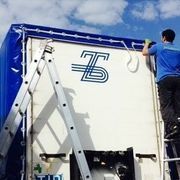 Uomo su una scala che ripara un camion bianco con un telo blu. Il camion ha il logo
