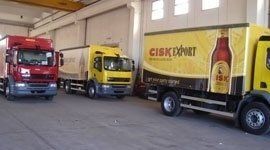 Tre camion per le consegne parcheggiati all'interno di un magazzino: uno rosso, due gialli e uno che pubblicizza la birra Cisk.