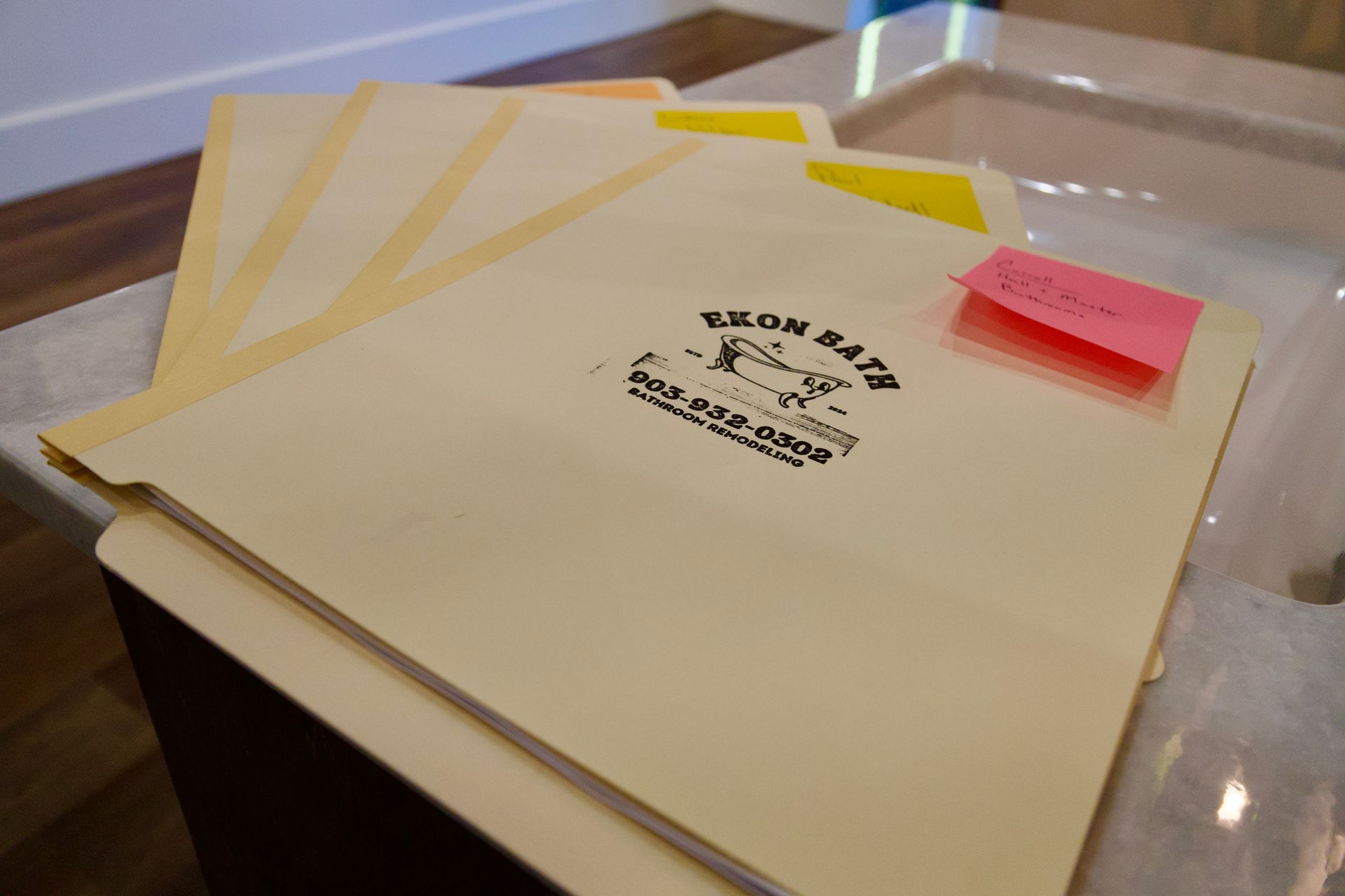 Cream-colored Ekon Bath folders on a desk, one labeled with a pink sticky note and yellow tabs.
