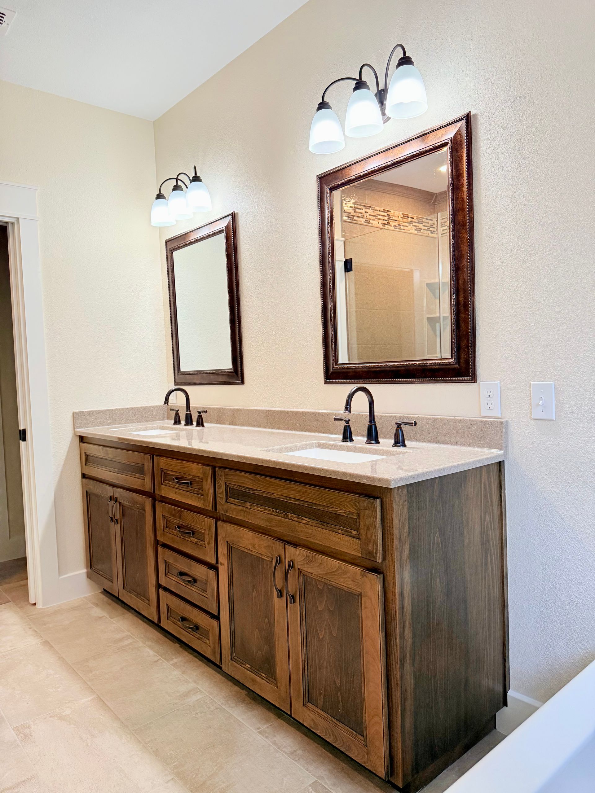 Bathroom with double vanity, two mirrors, and three-light sconces. Wooden cabinets, beige countertop, and light-colored walls.