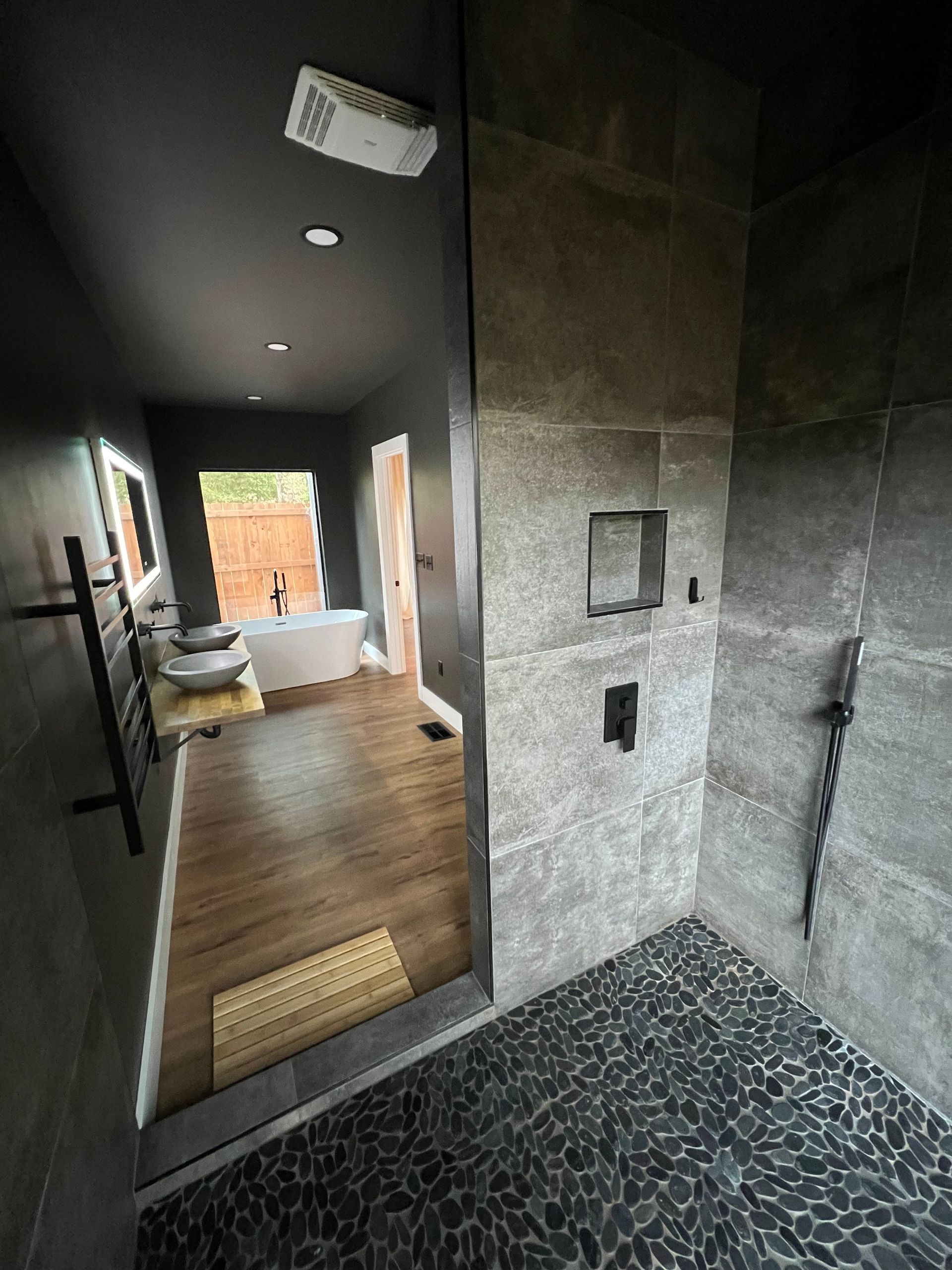 Modern bathroom with gray tiled shower and pebble floor, leading to a wooden floor and a white bathtub.