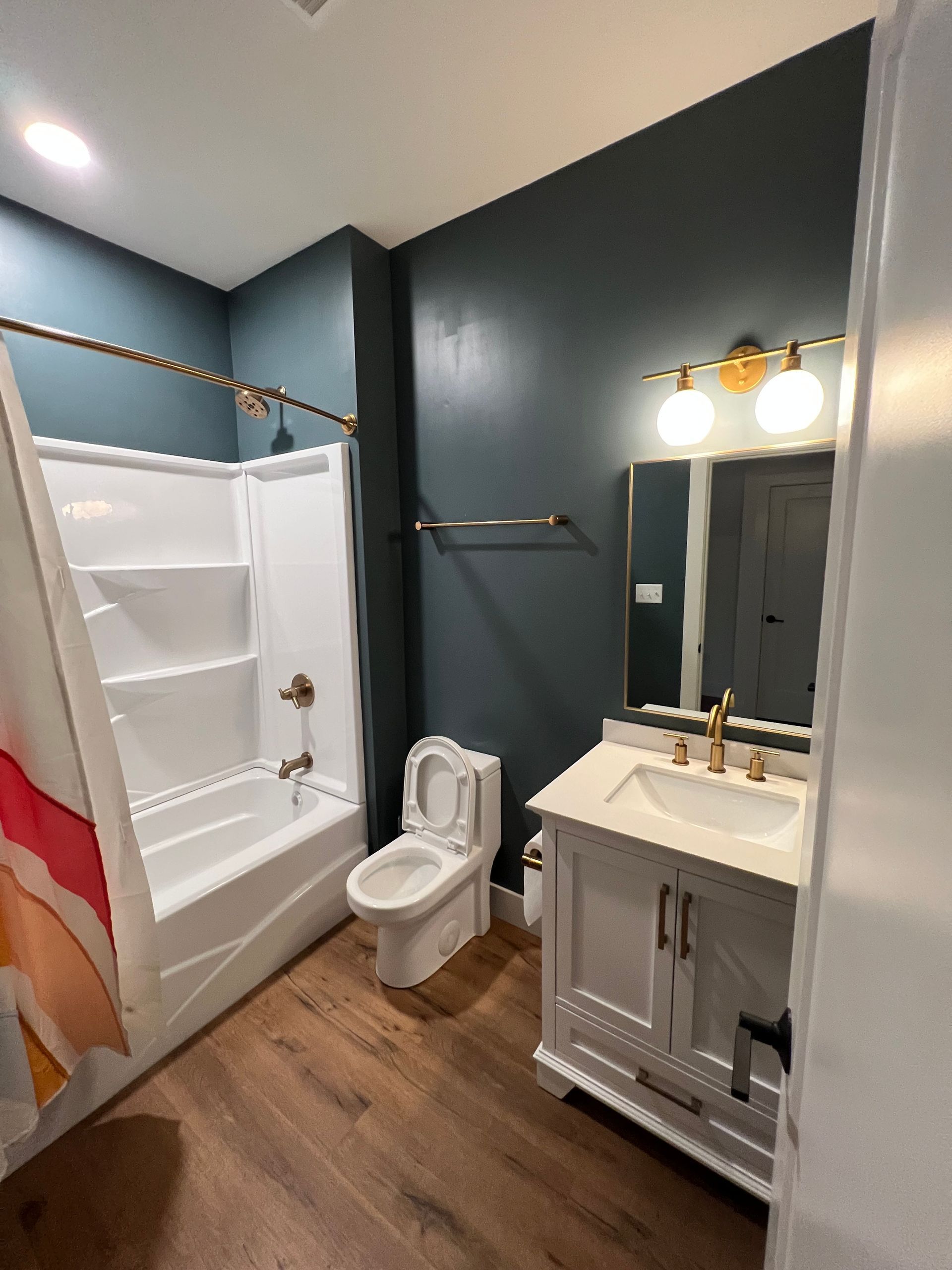 Bathroom with teal walls, white vanity, toilet, tub, gold fixtures, and wood-look flooring.