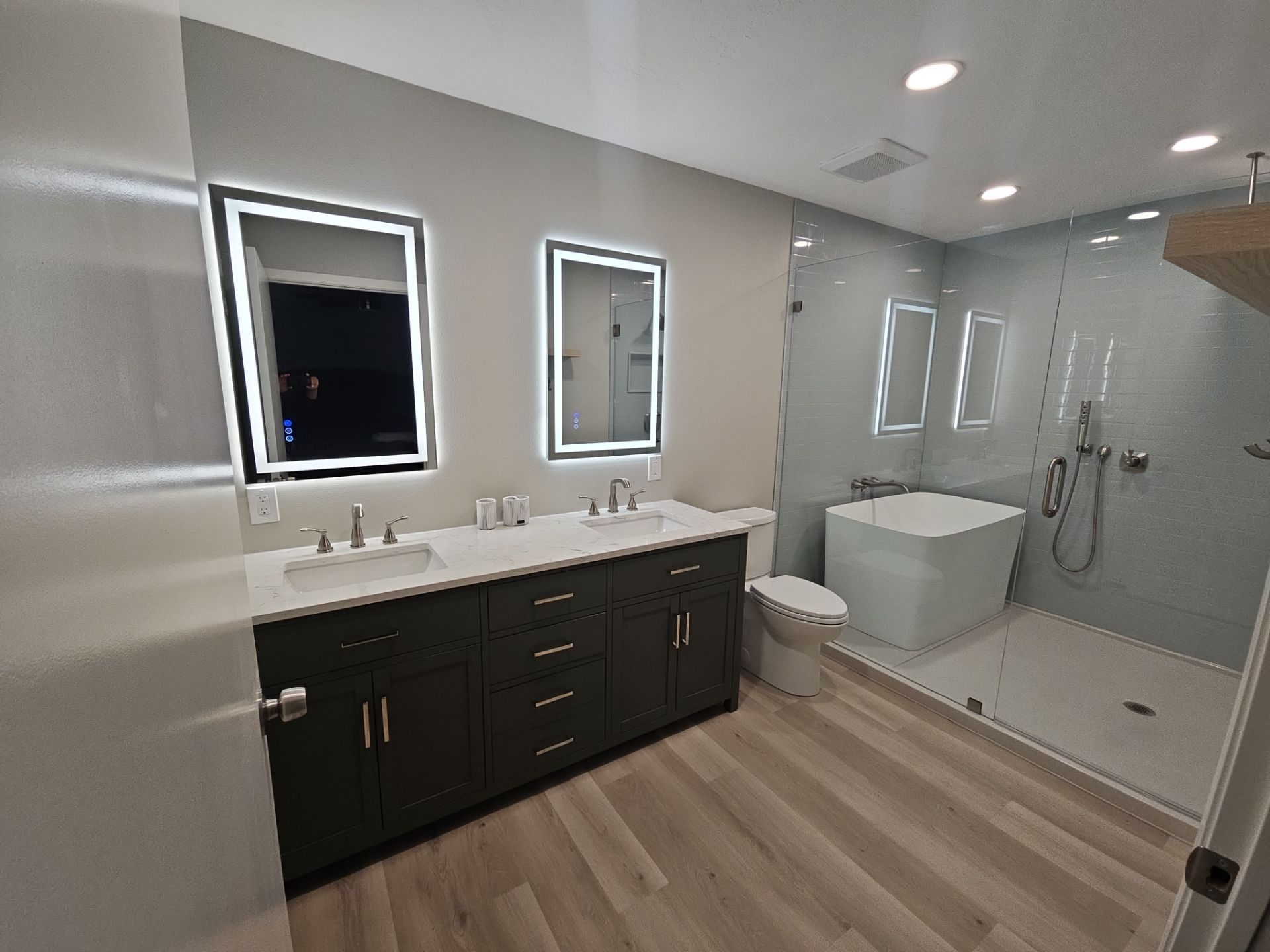 Modern bathroom with a double vanity, dark cabinets, two illuminated mirrors, and a glass-enclosed shower with a tub.