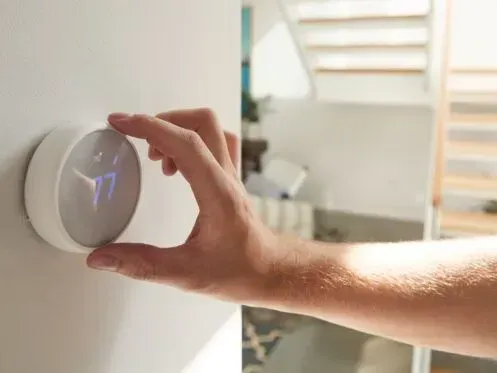 A person adjusting a round, white smart thermostat mounted on a wall in a brightly lit room.