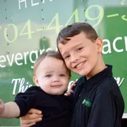 An older child holds a smiling toddler in front of a green sign with white text.