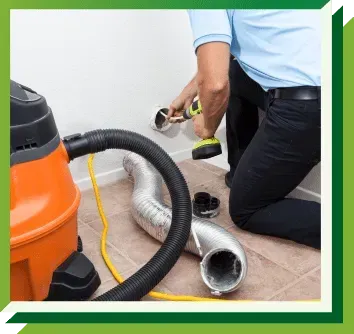 A person kneeling on the floor uses a power drill to service a wall vent near a vacuum and a disconnected dryer duct hose.