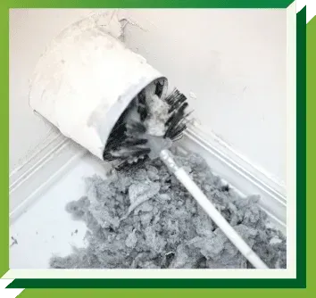 A dryer vent cleaning brush pulls a large clump of gray lint out of a white wall-mounted vent pipe.